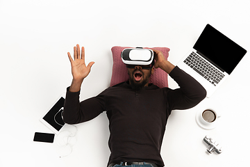 Image showing Emotional african-american man using VR-headset surrounded by gadgets isolated on white studio background, technologies. Emotional playing