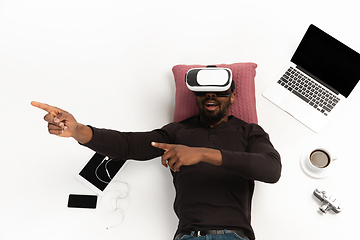 Image showing Emotional african-american man using VR-headset surrounded by gadgets isolated on white studio background, technologies. Emotional playing