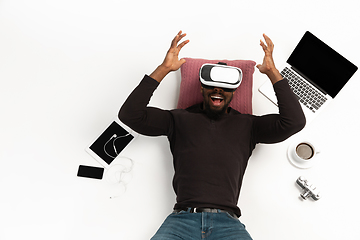 Image showing Emotional african-american man using VR-headset surrounded by gadgets isolated on white studio background, technologies. Emotional playing