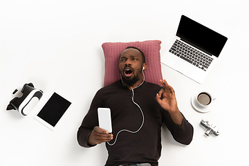 Image showing Emotional african-american man using phone surrounded by gadgets isolated on white studio background, technologies connecting people. Inspired singing