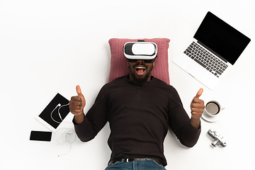 Image showing Emotional african-american man using VR-headset surrounded by gadgets isolated on white studio background, technologies. Thumbs up, happy