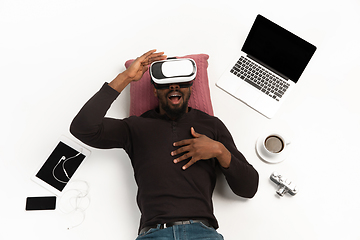 Image showing Emotional african-american man using VR-headset surrounded by gadgets isolated on white studio background, technologies. Emotional playing