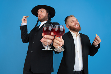 Image showing Portrait of a young orthodox jewish men isolated on blue studio background, meeting the Passover, eating Aman\'s ears with wine