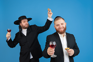 Image showing Portrait of a young orthodox jewish men isolated on blue studio background, meeting the Passover, eating Aman\'s ears with wine