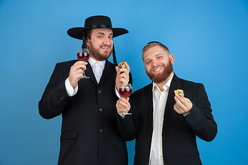 Image showing Portrait of a young orthodox jewish men isolated on blue studio background, meeting the Passover, eating Aman\'s ears with wine