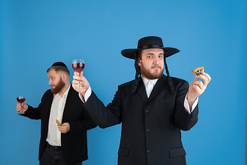 Image showing Portrait of a young orthodox jewish men isolated on blue studio background, meeting the Passover, eating Aman\'s ears with wine