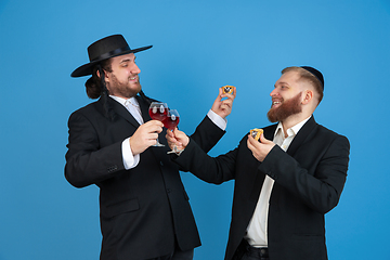 Image showing Portrait of a young orthodox jewish men isolated on blue studio background, meeting the Passover, eating Aman\'s ears with wine