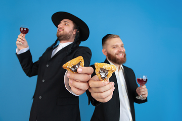 Image showing Portrait of a young orthodox jewish men isolated on blue studio background, meeting the Passover, eating Aman\'s ears with wine