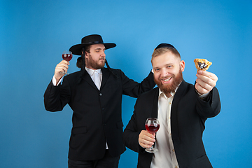 Image showing Portrait of a young orthodox jewish men isolated on blue studio background, meeting the Passover, eating Aman\'s ears with wine