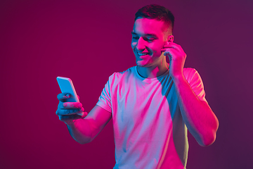 Image showing Caucasian man\'s portrait isolated on pink-purple studio background in neon light, with smartphone and earphones