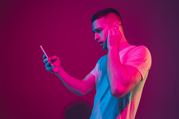 Image showing Caucasian man\'s portrait isolated on pink-purple studio background in neon light, with smartphone and earphones