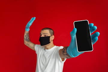 Image showing Caucasian young man in protective face mask and medical gloves on red studio background - showing blank phone\'s screen