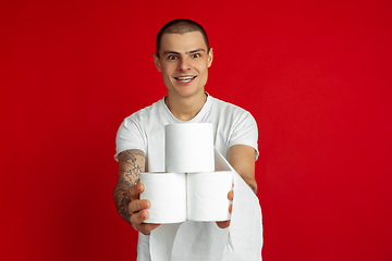 Image showing Caucasian young man\'s portrait on red studio background - holding toilet papers, essential goods during quarantine and self-insulation