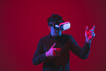 Image showing Caucasian man\'s portrait isolated on red studio background in purple-blue neon light, emotional and beautiful, playing wore VR-headset