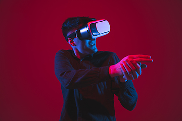 Image showing Caucasian man\'s portrait isolated on red studio background in purple-blue neon light, emotional and beautiful, playing wore VR-headset