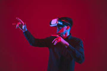 Image showing Caucasian man\'s portrait isolated on red studio background in purple-blue neon light, emotional and beautiful, playing wore VR-headset