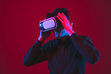 Image showing Caucasian man\'s portrait isolated on red studio background in purple-blue neon light, emotional and beautiful, playing wore VR-headset