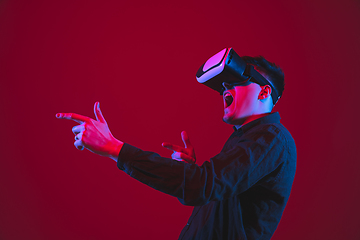 Image showing Caucasian man\'s portrait isolated on red studio background in purple-blue neon light, emotional and beautiful, playing wore VR-headset