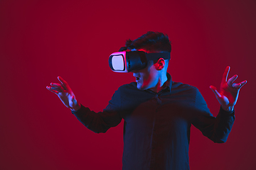 Image showing Caucasian man\'s portrait isolated on red studio background in purple-blue neon light, emotional and beautiful, playing wore VR-headset