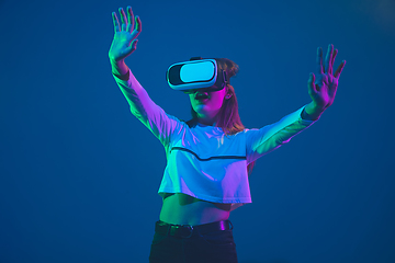 Image showing Caucasian woman\'s portrait isolated on blue studio background in pink-green neon light, stylish and beautiful, playing with VR-headset