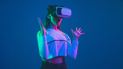 Image showing Caucasian woman\'s portrait isolated on blue studio background in pink-green neon light, stylish and beautiful, playing with VR-headset