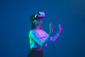 Image showing Caucasian woman\'s portrait isolated on blue studio background in pink-green neon light, stylish and beautiful, playing with VR-headset