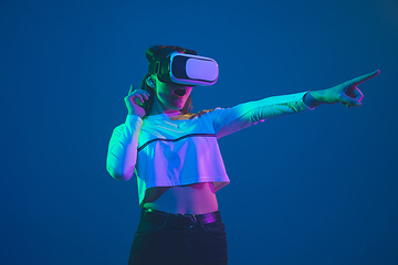 Image showing Caucasian woman\'s portrait isolated on blue studio background in pink-green neon light, stylish and beautiful, playing with VR-headset