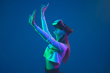 Image showing Caucasian woman\'s portrait isolated on blue studio background in pink-green neon light, stylish and beautiful, playing with VR-headset