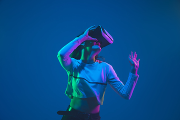 Image showing Caucasian woman\'s portrait isolated on blue studio background in pink-green neon light, stylish and beautiful, playing with VR-headset