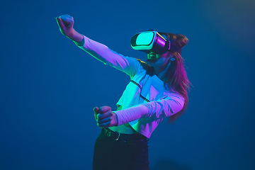 Image showing Caucasian woman\'s portrait isolated on blue studio background in pink-green neon light, stylish and beautiful, playing with VR-headset