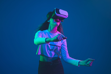 Image showing Caucasian woman\'s portrait isolated on blue studio background in pink-green neon light, stylish and beautiful, playing with VR-headset