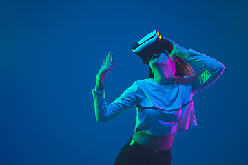 Image showing Caucasian woman\'s portrait isolated on blue studio background in pink-green neon light, stylish and beautiful, playing with VR-headset