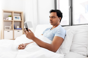 Image showing indian man in earphones with phone in bed at home