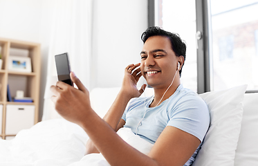 Image showing indian man in earphones with phone in bed at home