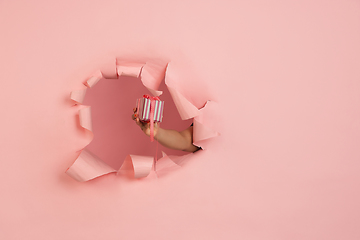 Image showing Female hand giving gift in torn coral, pink paper hole background, celebration