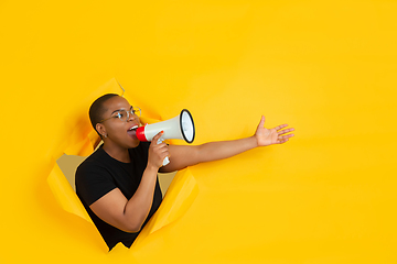 Image showing Cheerful young woman poses in torn yellow paper hole background, emotional and expressive, shouting and calling with speaker