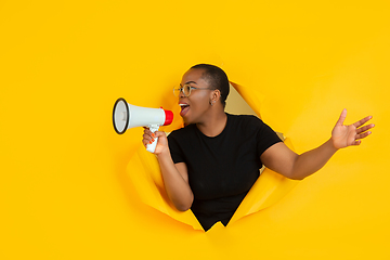 Image showing Cheerful young woman poses in torn yellow paper hole background, emotional and expressive, shouting and calling with speaker