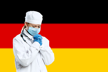 Image showing Female young doctor with stethoscope and face mask praying for God with Germany national flag on background