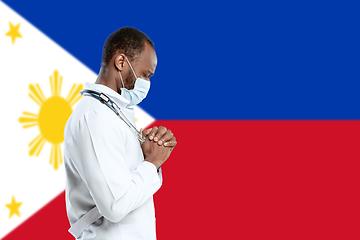 Image showing Young doctor with stethoscope and face mask praying for God with Philippines national flag on background