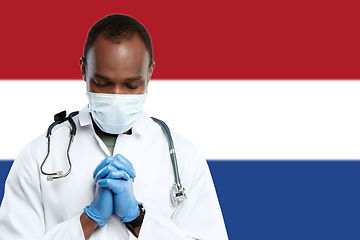 Image showing Young doctor with stethoscope and face mask praying for God with Netherlands national flag on background