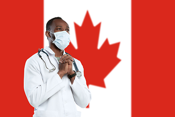 Image showing Young doctor with stethoscope and face mask praying for God with Canada national flag on background