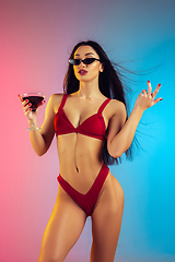 Image showing Fashion portrait of young fit and sportive woman with cocktail in stylish red luxury swimwear on gradient background. Perfect body ready for summertime.