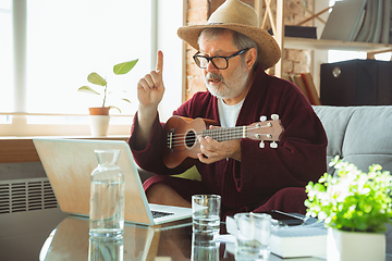 Image showing Mature senior older man during quarantine, realizing how important stay at home during virus outbreak, giving concert of taking online lessons of guitar playing