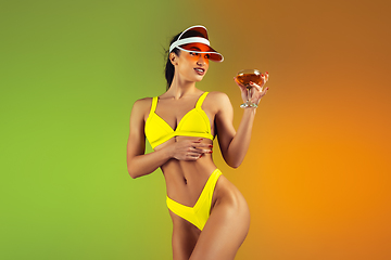 Image showing Fashion portrait of young fit and sportive woman with cocktail in stylish yellow luxury swimwear on gradient background. Perfect body ready for summertime.