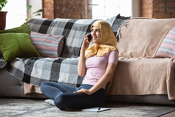 Image showing A pretty young muslim woman at home during quarantine and self-insulation, using smartphone for talking, online lessons, shopping