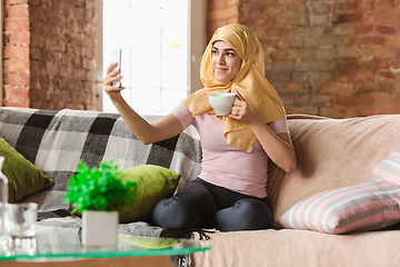 Image showing A pretty young muslim woman at home during quarantine and self-insulation, using smartphone for selfie or videocall, online lessons, shopping