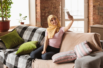 Image showing A pretty young muslim woman at home during quarantine and self-insulation, using smartphone for selfie or videocall, online lessons, shopping