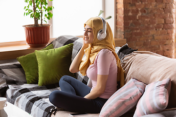 Image showing A pretty young muslim woman at home during quarantine and self-insulation, using headphones, listen to music, enjoying