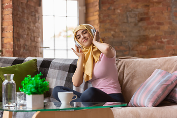 Image showing A pretty young muslim woman at home during quarantine and self-insulation, using headphones, listen to music, enjoying