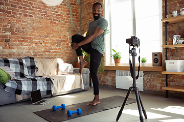 Image showing Young african-american man teaching at home online courses of fitness, aerobic, sporty lifestyle during quarantine, reording on camera, streaming